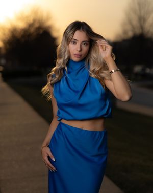 A woman in a flowing blue dress poses dramatically at sunset in an outdoor setting.
