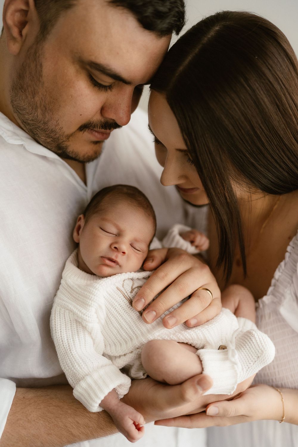 A couple in white clothing tenderly cradles their sleeping newborn baby, sharing an intimate family portrait moment.