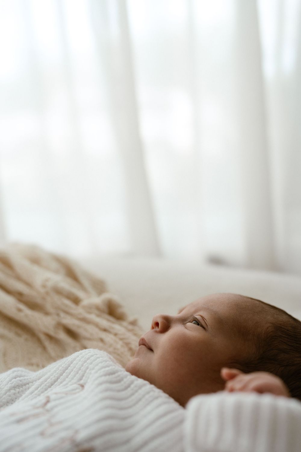 A peaceful newborn baby sleeping wrapped in a white knit blanket with soft natural light in the background.