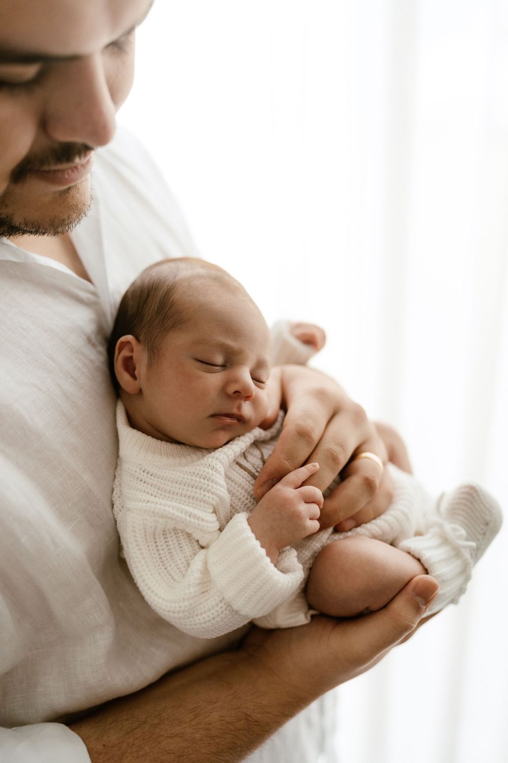 A father tenderly cradles his sleeping newborn baby dressed in white knit outfit, sharing a peaceful bonding moment.