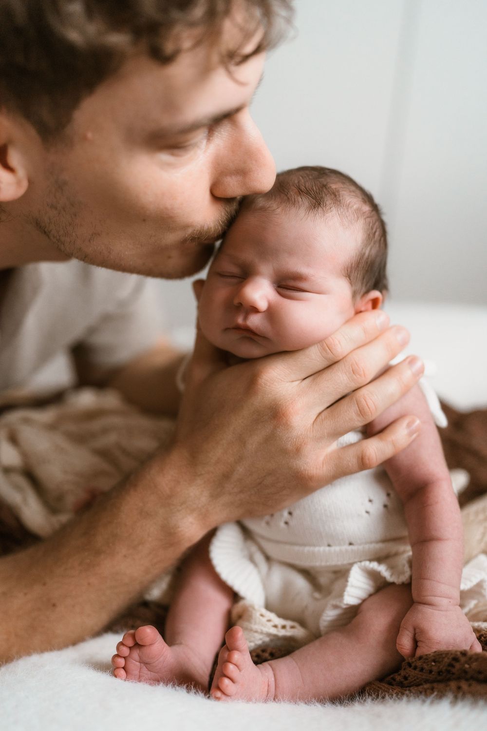 A loving father gently kisses his newborn baby while cradling the infant in his hands on a soft white blanket.