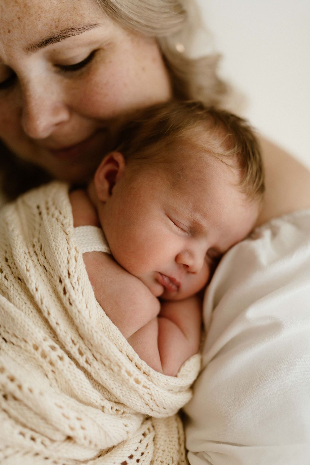 A mother tenderly holds her sleeping newborn baby wrapped in a soft cream knit blanket.