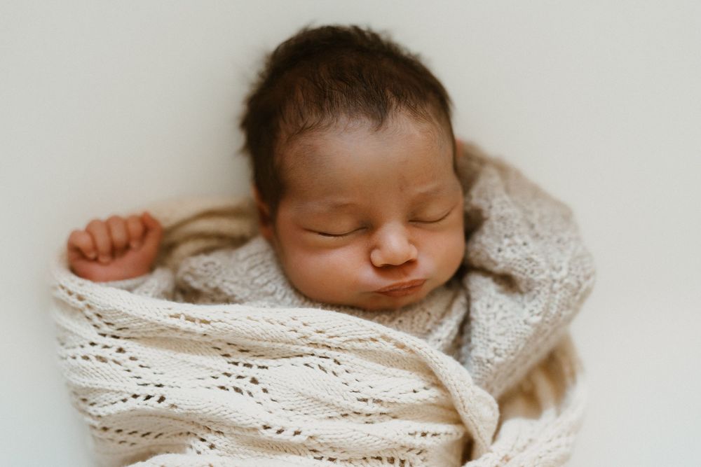 Peaceful newborn baby swaddled in a soft cream knit wrap, sleeping soundly with dark hair and tiny fist visible.
