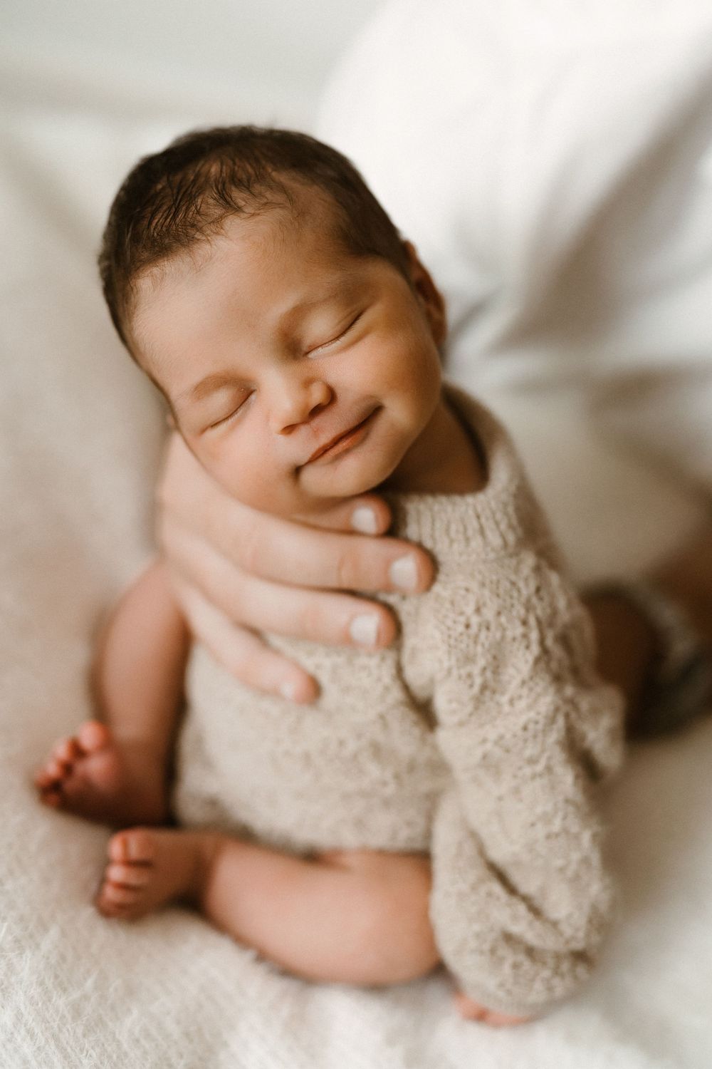 Smiling newborn baby in beige knit romper cradled gently by parent hands on white blanket.