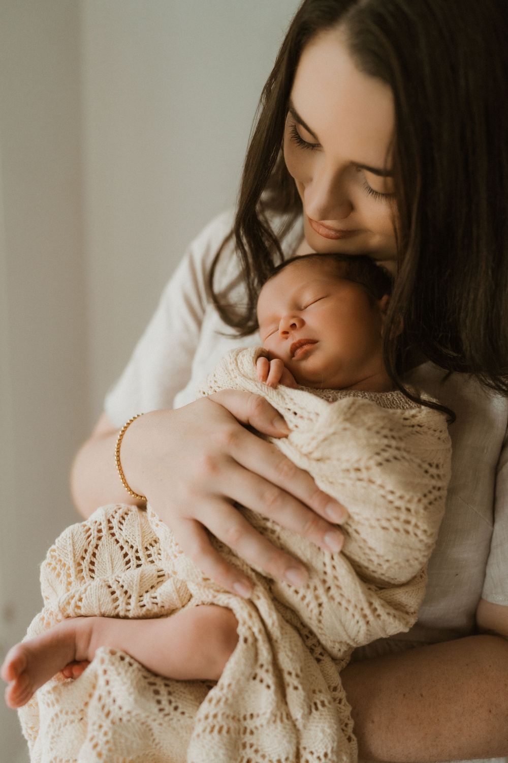 A mother with dark hair tenderly holds her sleeping newborn wrapped in a cream knit blanket.