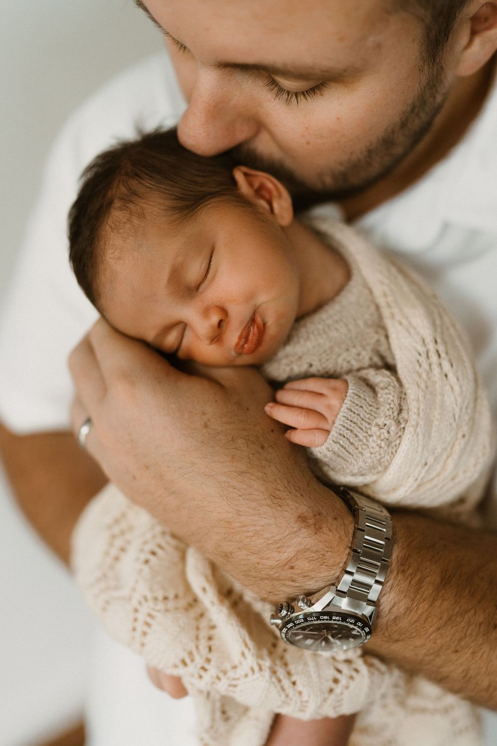A bearded father tenderly kisses his sleeping newborn baby wrapped in a cozy knit blanket while wearing a silver watch.