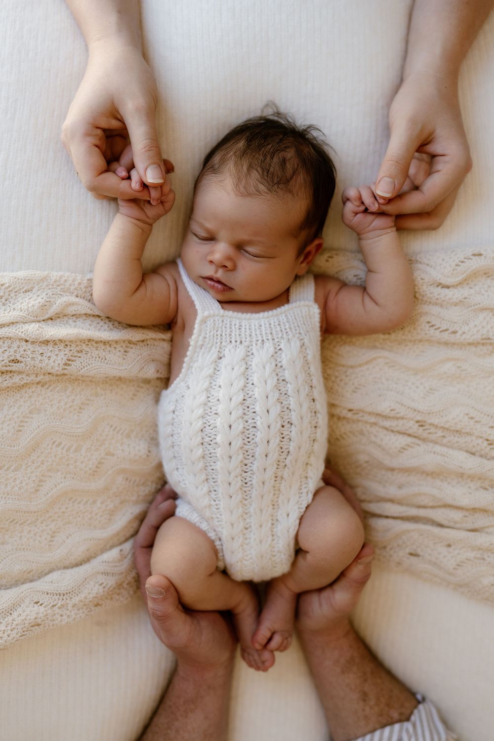 Sleeping newborn baby in white knit romper held gently by parents hands on a soft cream blanket.