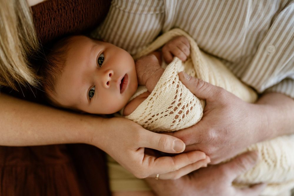 Newborn baby wrapped in cream knit blanket cradled gently in parents hands, peaceful infant portrait photography.