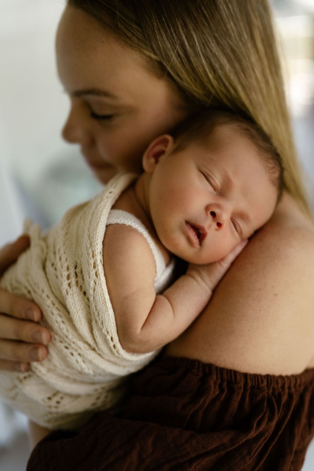 A mother with blonde hair gently holds her sleeping newborn baby wrapped in a cream knit swaddle against her shoulder.