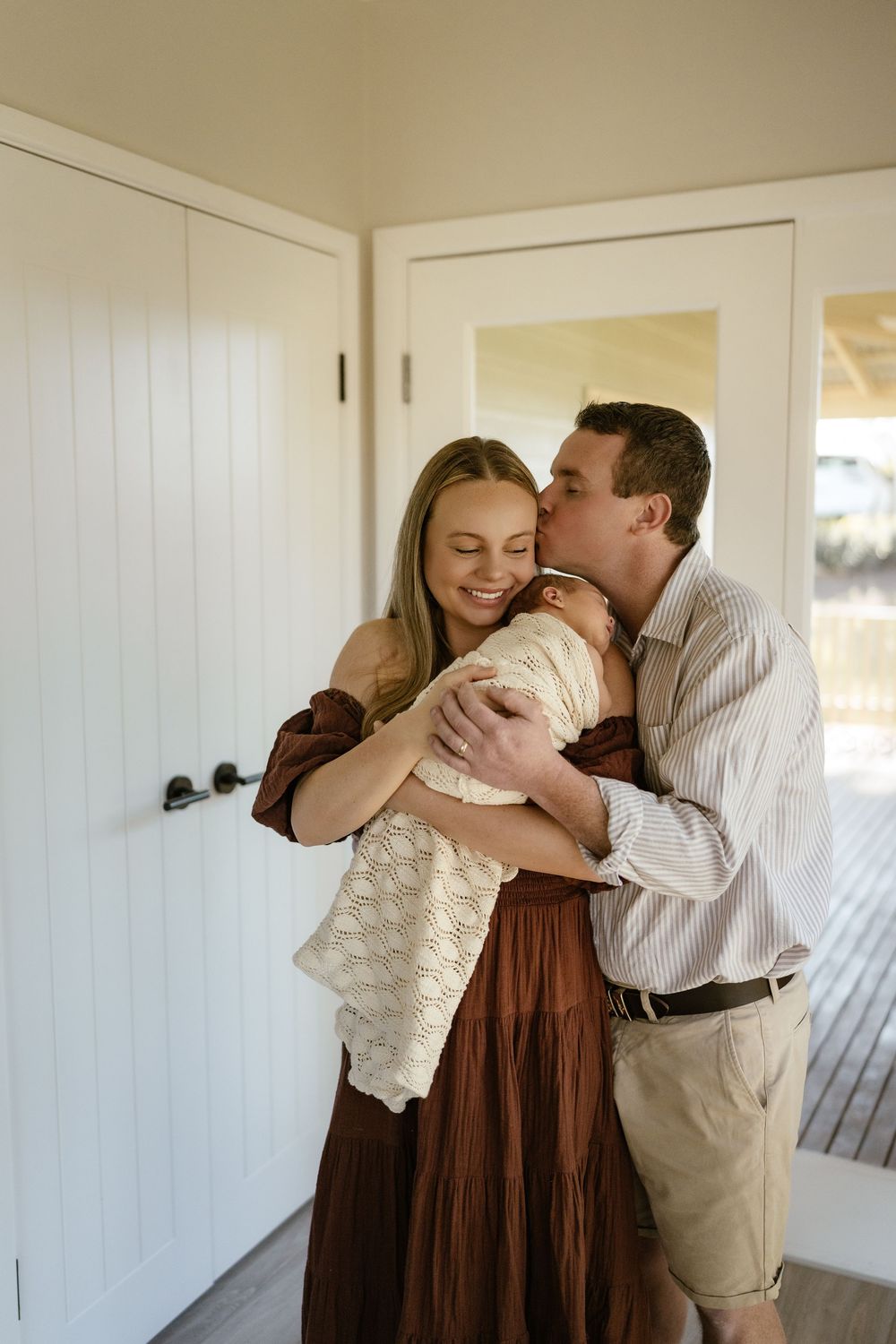 A couple shares a tender moment, kissing their newborn baby wrapped in a white blanket inside a bright home.