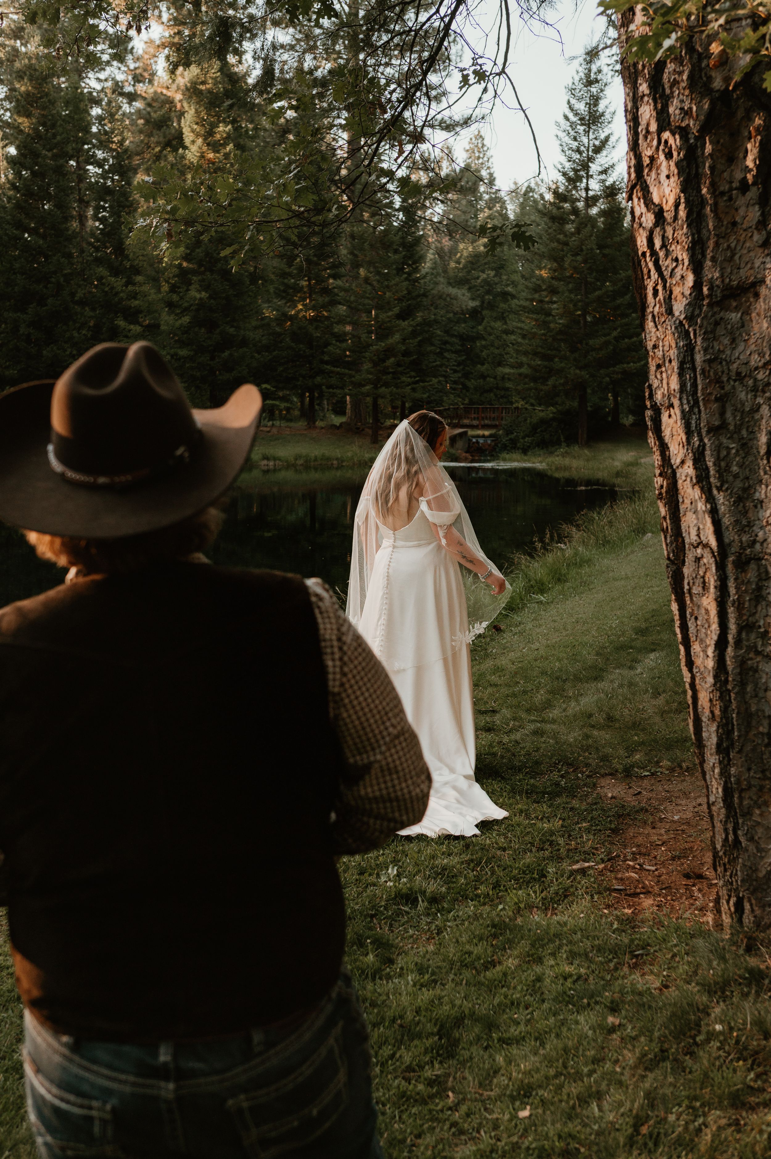 A romantic forest scene with a flowing white dress and cowboy hat silhouette among tall pine trees at sunset.