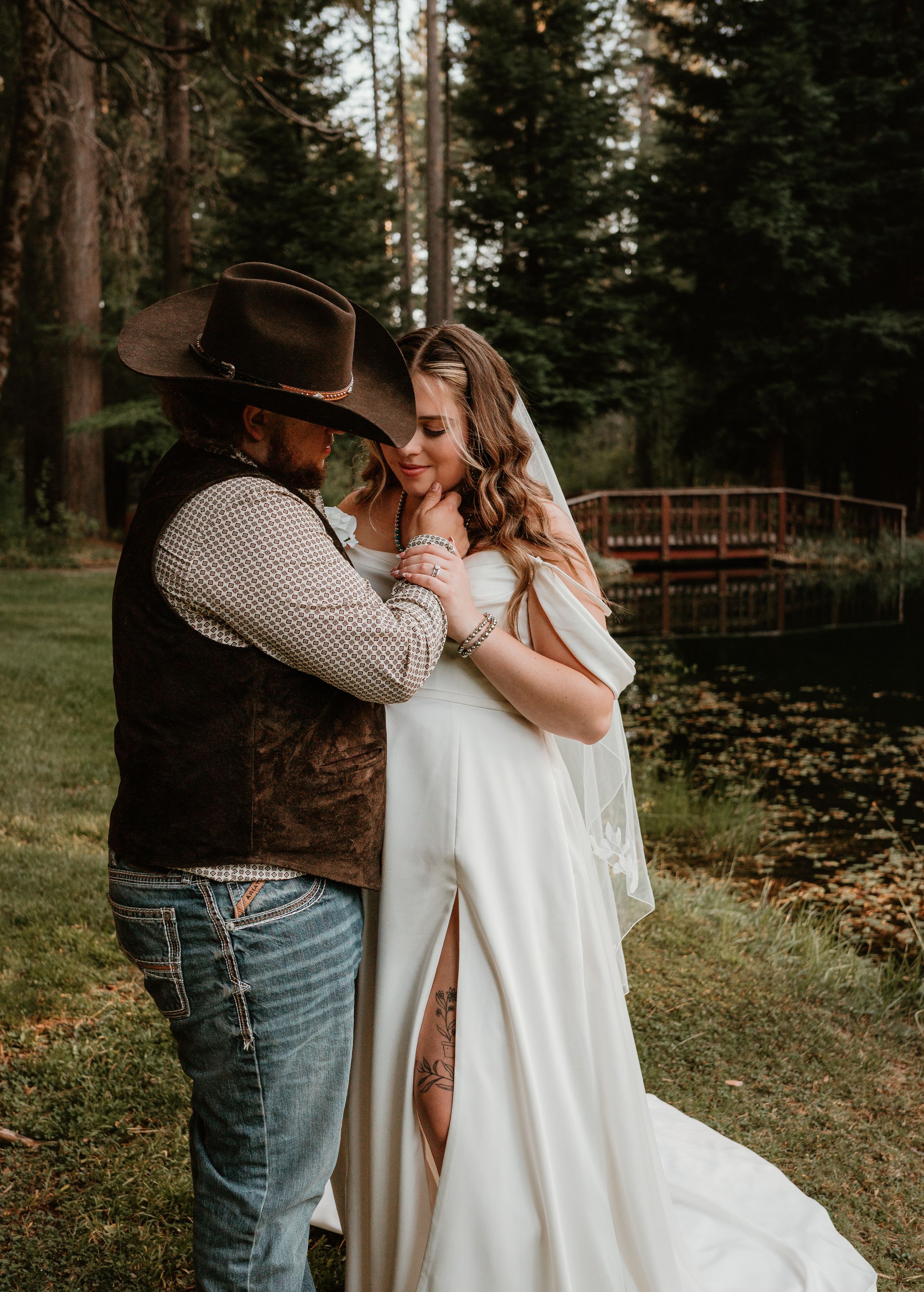 A couple embraces in a rustic woodland setting with pine trees and a stone wall in the background.
