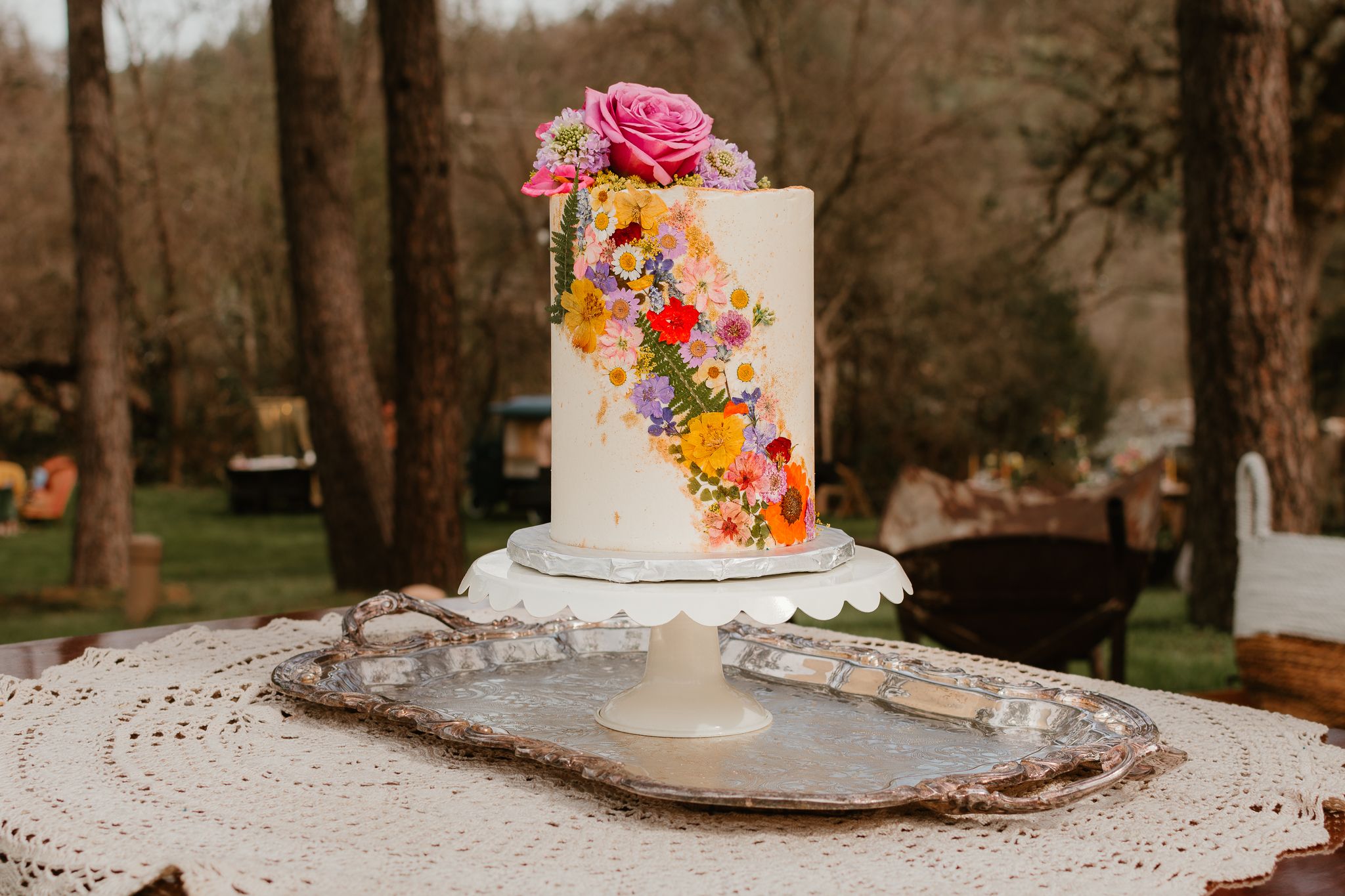 White wedding cake decorated with colorful fresh flowers displayed in an outdoor setting.