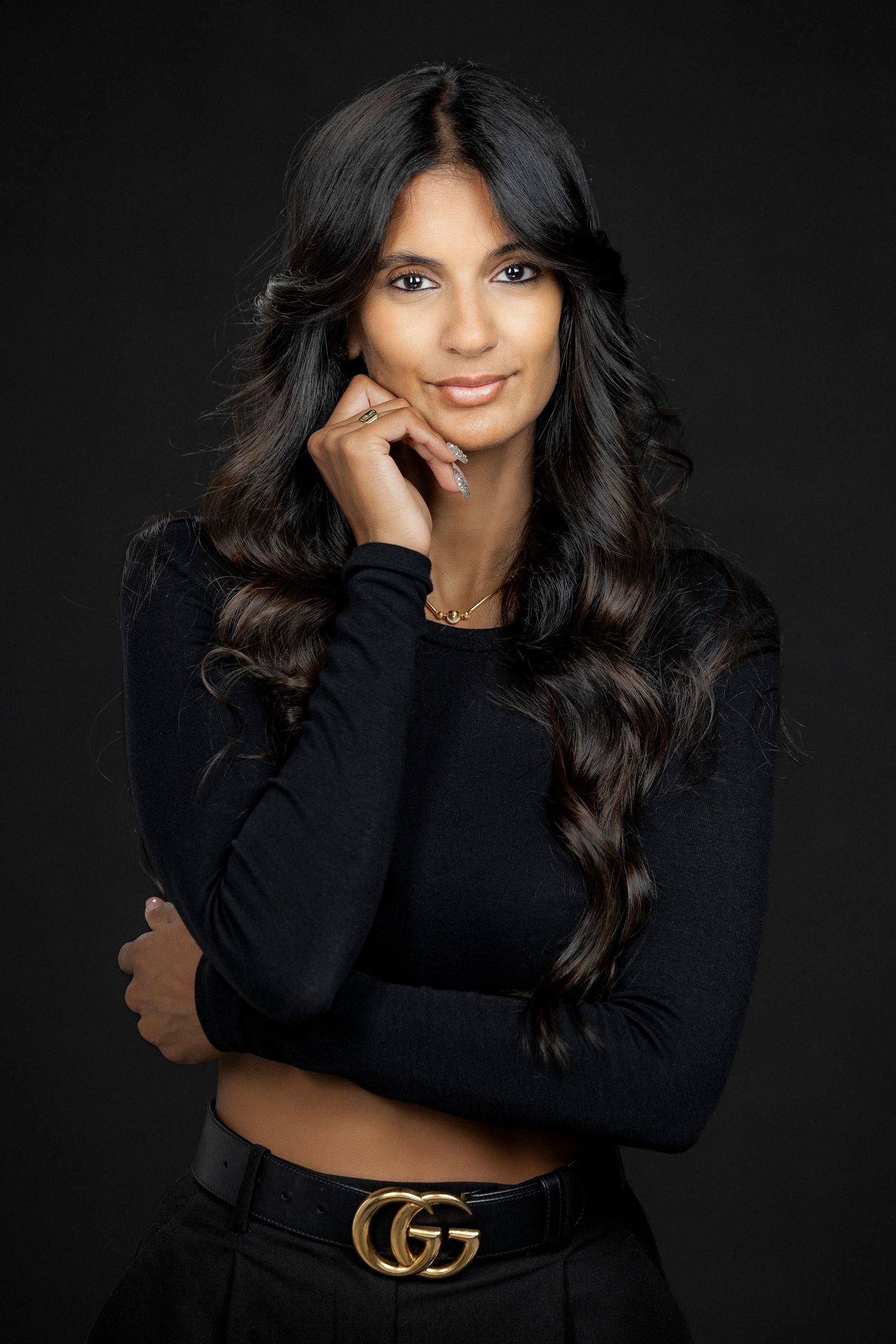 Portrait of a person in black long sleeve top with long dark wavy hair and a designer belt against dark background.
