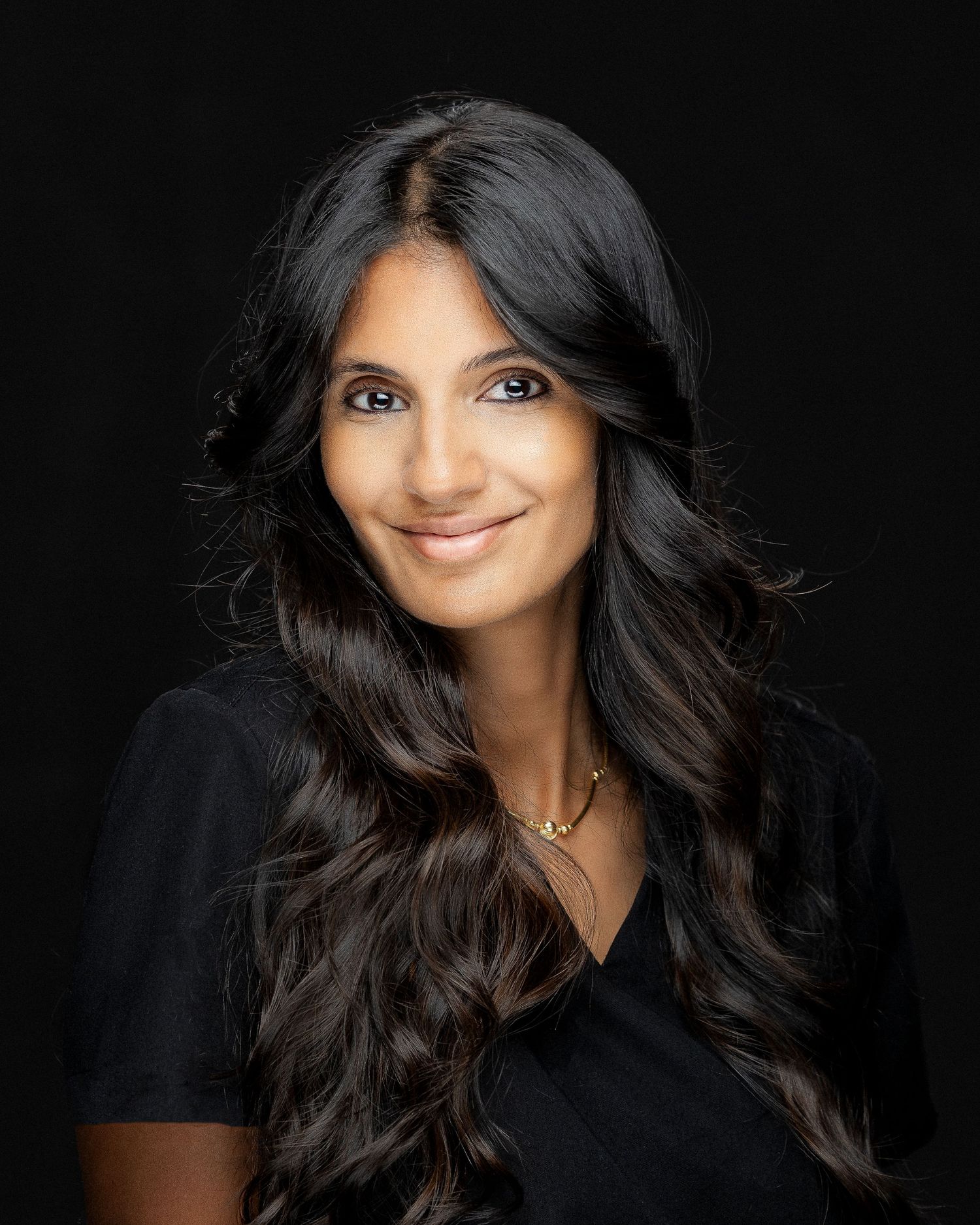 A woman in a black top smiles warmly against a dark background with graceful head tilt and flowing dark hair.