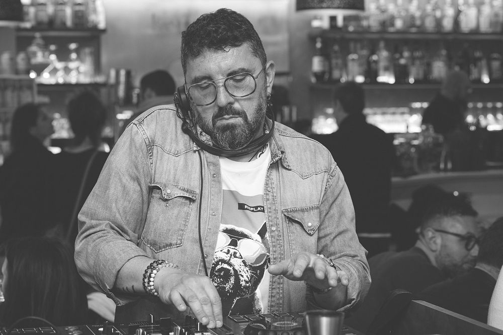 Bartender wearing denim and glasses mixing drinks at a bar counter in black and white.