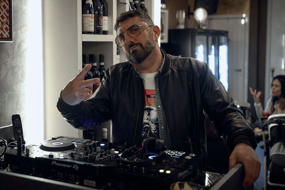 DJ in leather jacket mixing music at turntable setup in dimly lit venue.