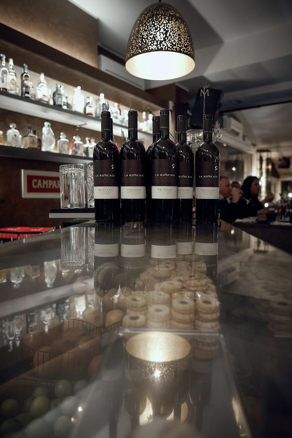 Wine bottles lined up on reflective bar counter under decorative pendant light.