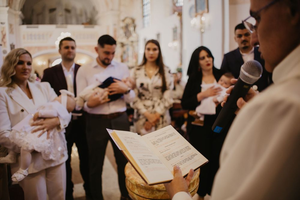 People gathered in a church for a baptismal ceremony as a priest reads from a book.
