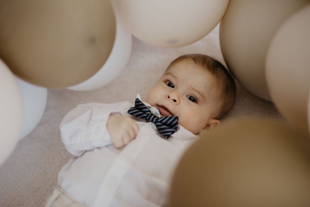 Baby in white outfit with bowtie surrounded by white balloons.