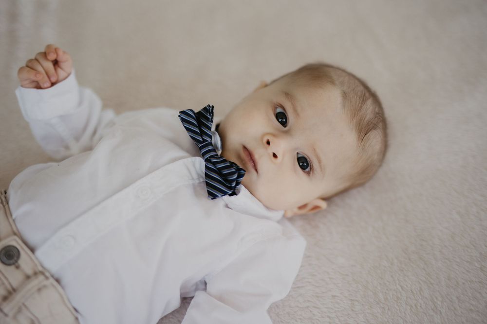 Baby in white outfit wearing blue striped bowtie lying on light colored surface.