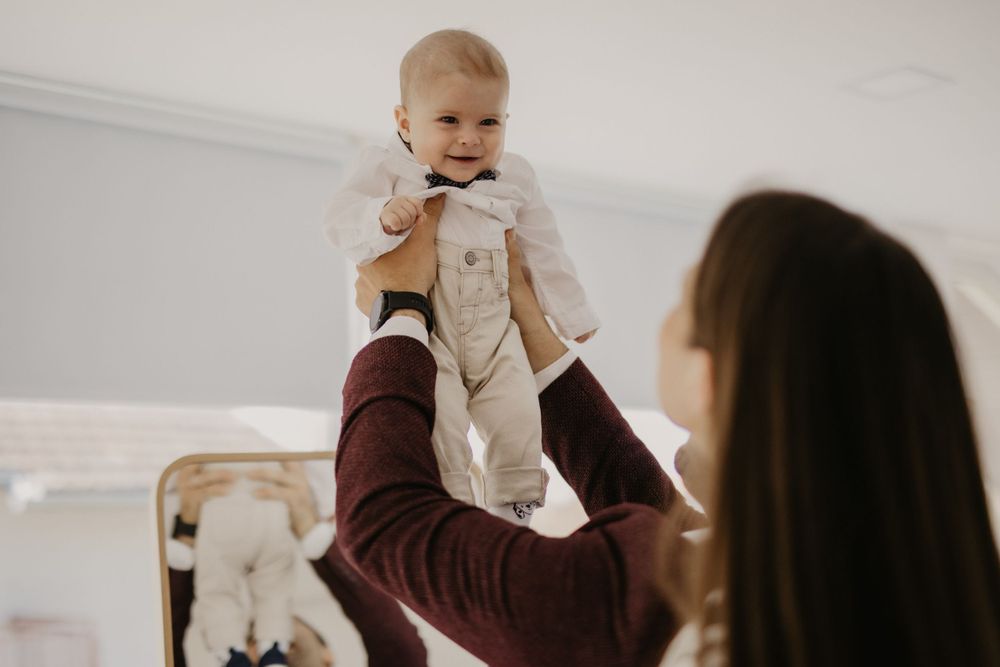 Parent lifting baby wearing formal outfit in front of mirror.
