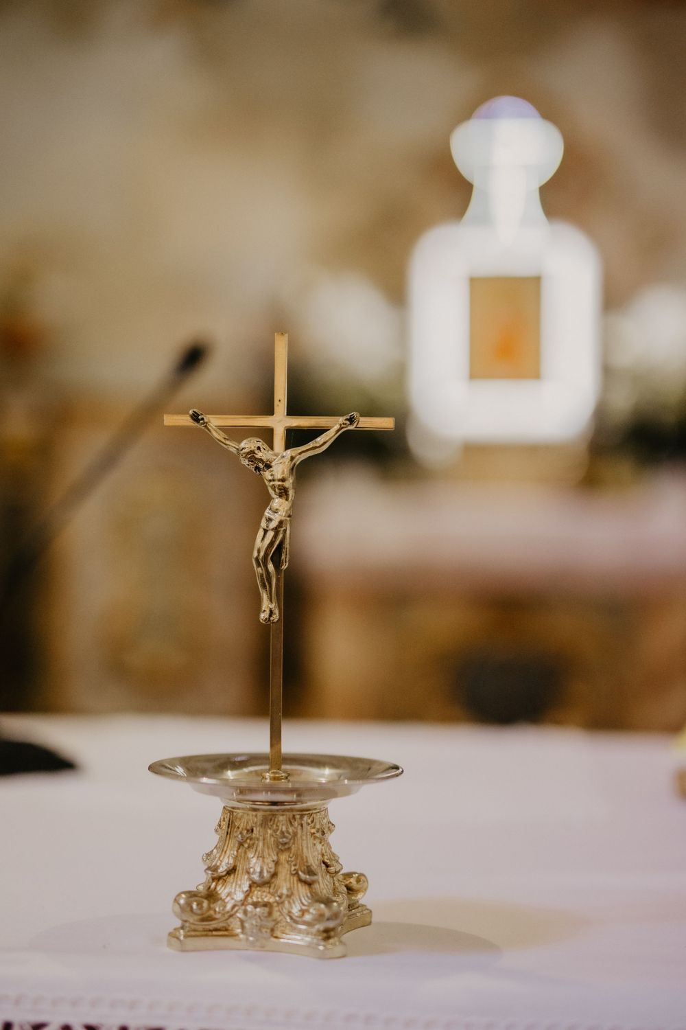 Golden crucifix displayed on an ornate metal stand with a blurred white figure in the background.
