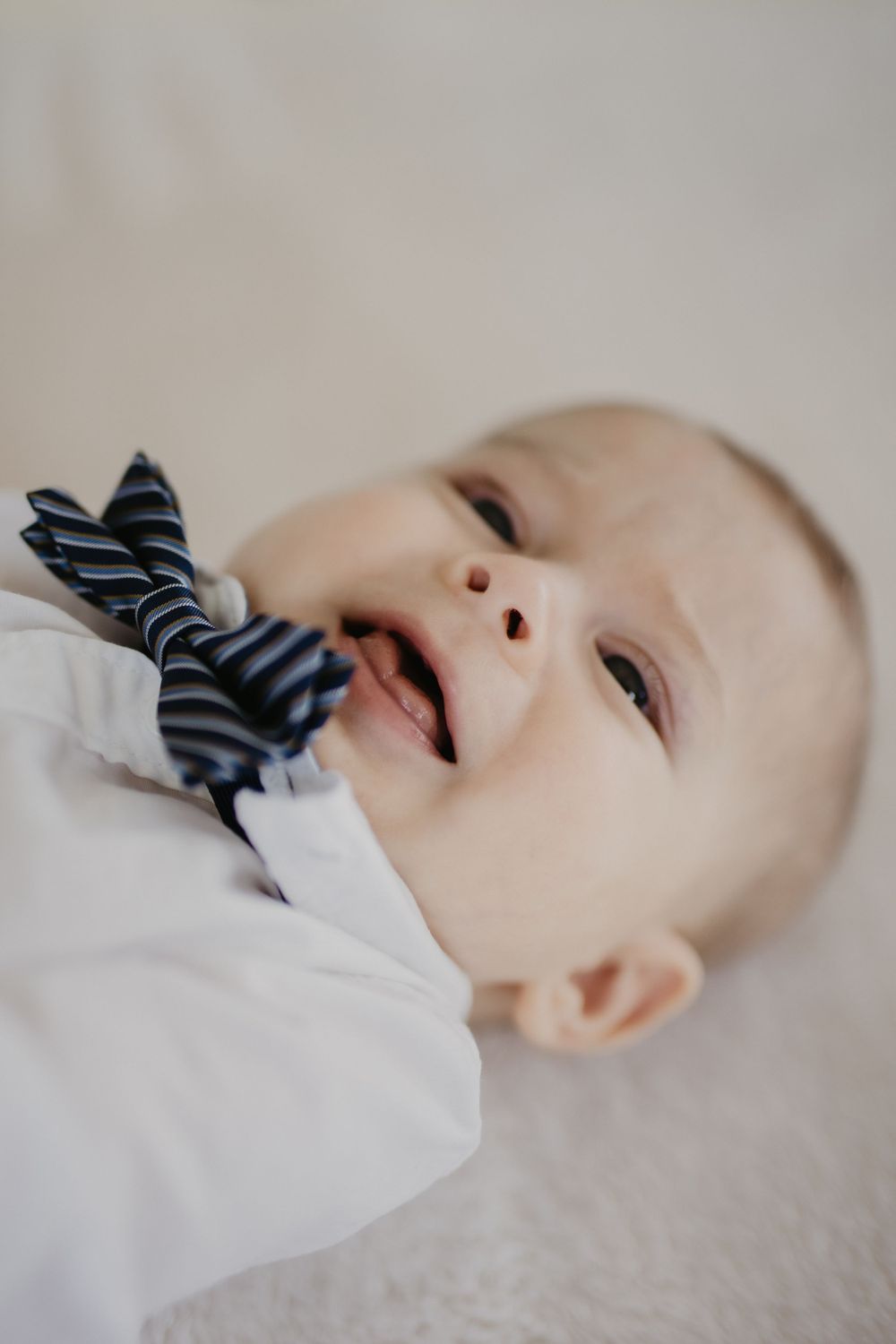 Smiling baby wearing striped bowtie and white outfit laying down.