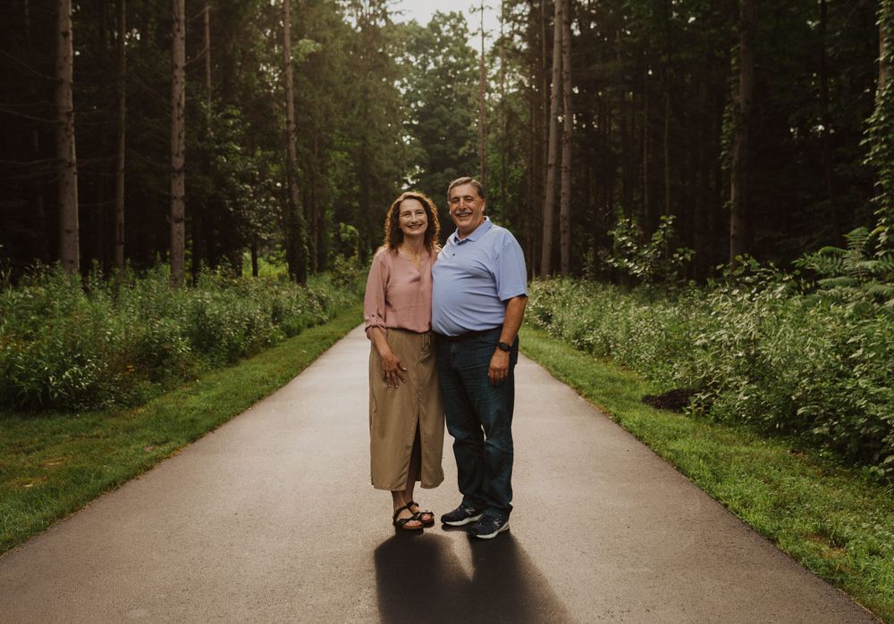A couple poses together on a forest path wearing casual attire.