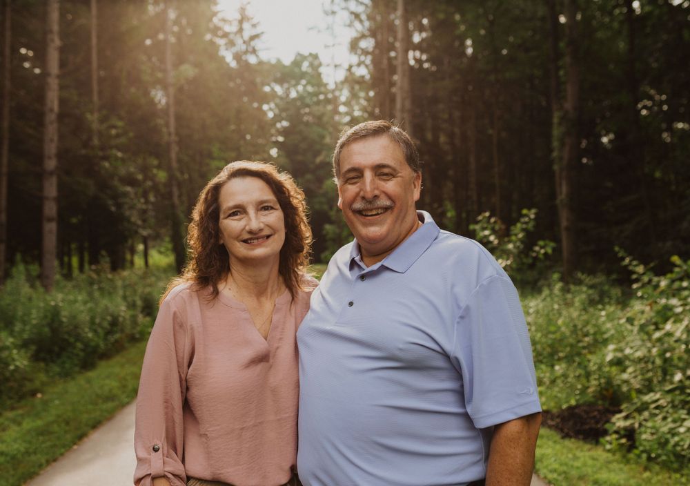 A couple stands together on a wooded path during sunset.