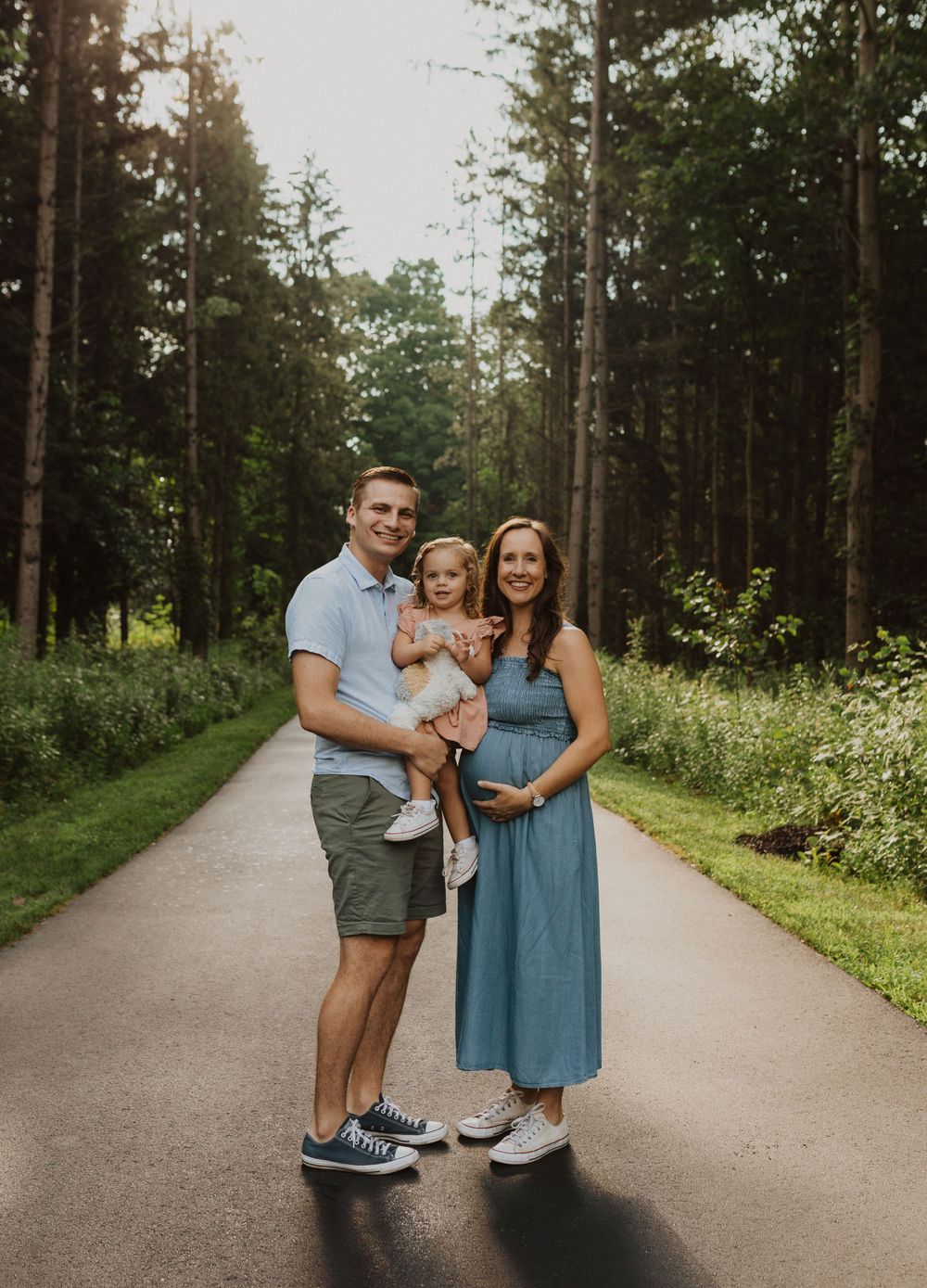 A family poses on a tree-lined path in a forest setting, with the parent wearing a blue dress showing a baby bump.