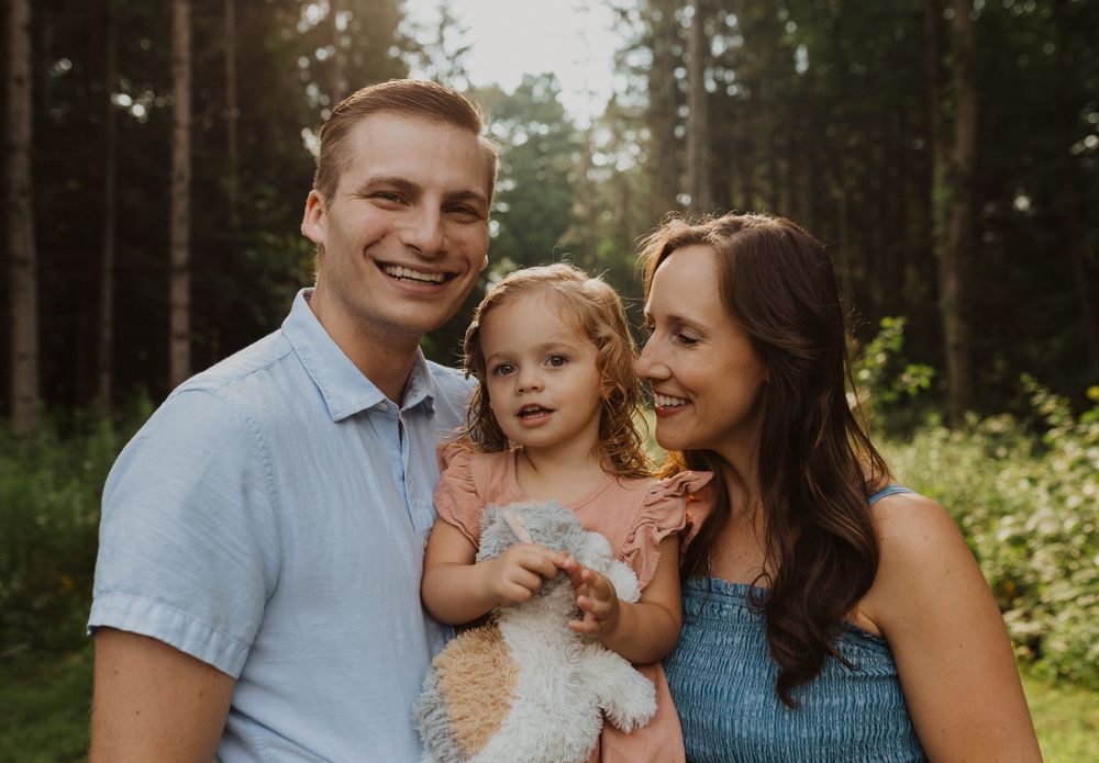 A close-up portrait of a family of three smiling together in a wooded setting.