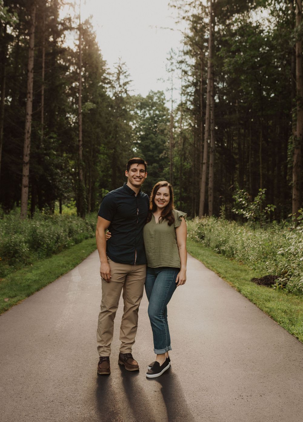 A couple stands together smiling on a paved forest path during golden hour.