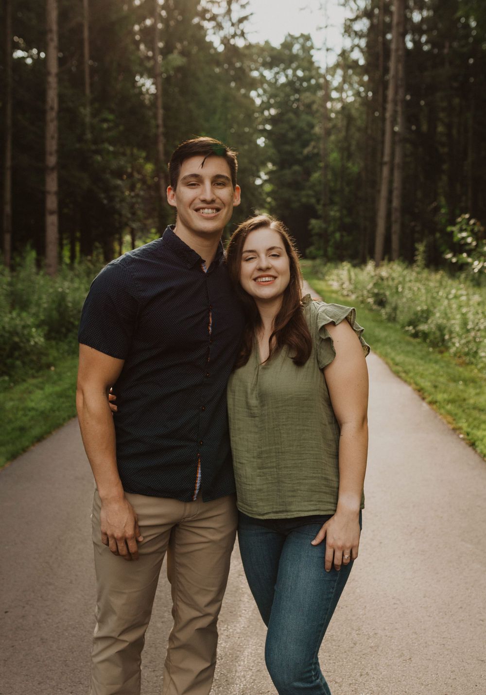 A couple embraces and smiles while standing on a paved trail surrounded by tall evergreen trees.