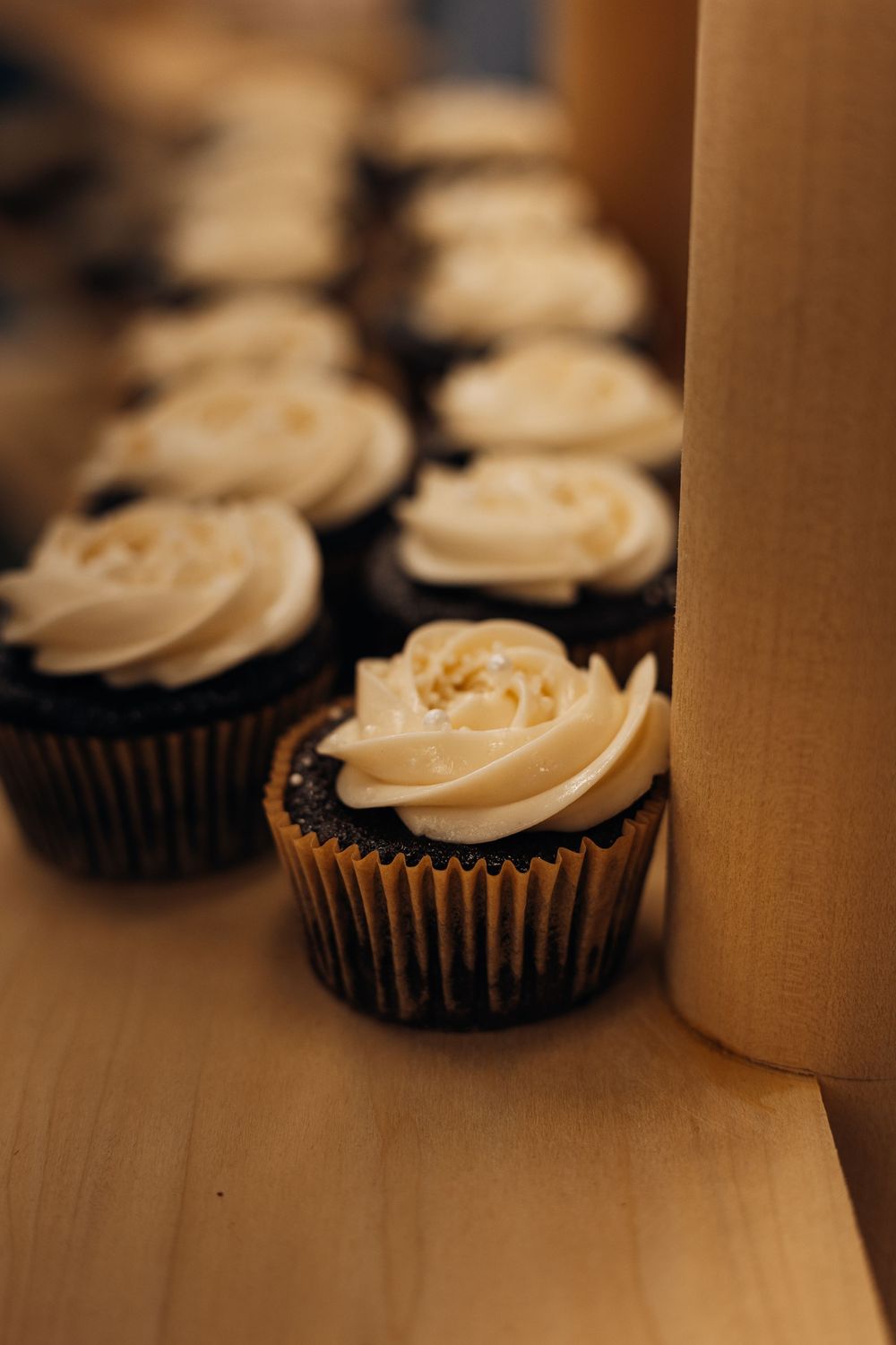 Rows of chocolate cupcakes with swirled vanilla buttercream frosting displayed on a wooden surface.