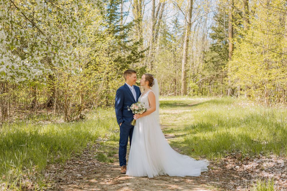 A bride and groom share a romantic moment on a sunlit forest path surrounded by lush green spring trees.