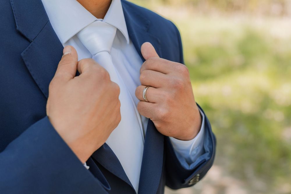 Groom in navy suit adjusting silver tie, wearing gold wedding ring, outdoor wedding preparation close-up.
