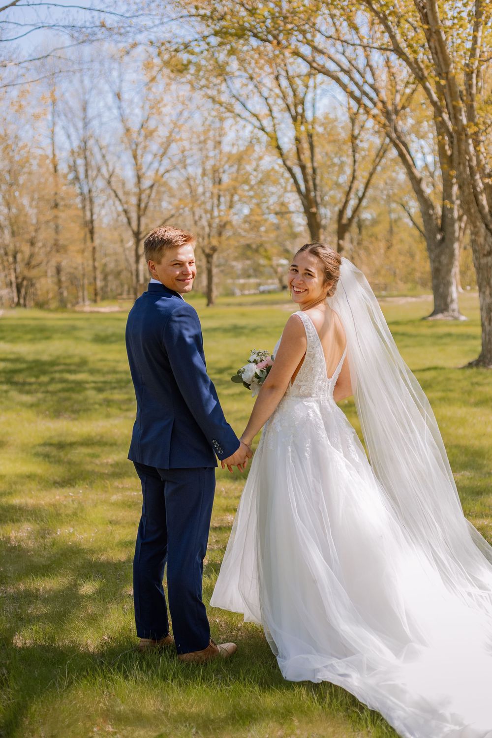 Bride and groom holding hands in a sunny park, smiling back at camera, wearing white gown with veil and navy suit.