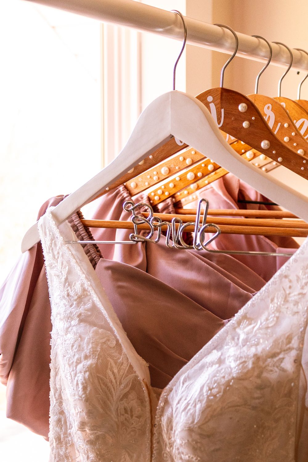 Wedding dress and bridesmaid gowns hanging on wooden hangers, including a decorative Bride hanger with pearl accents.