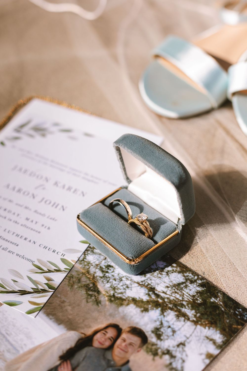 Gold diamond engagement ring in teal velvet box beside wedding invitation and couple photo.