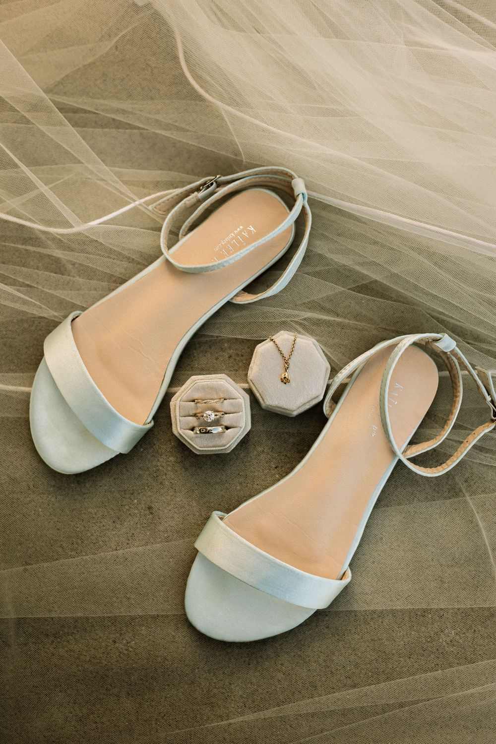 Elegant white bridal sandals with ankle straps beside a velvet ring box with wedding rings and gold necklace on gray surface.