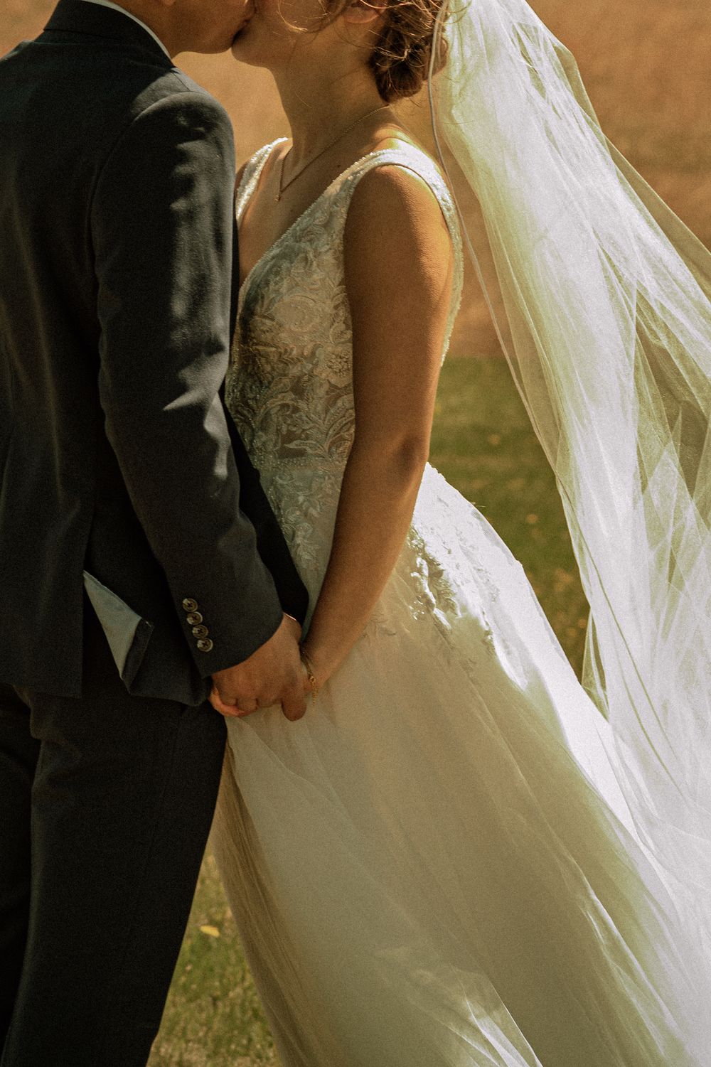 A bride and groom share a kiss while holding hands, her lace gown and flowing veil catching sunlight.