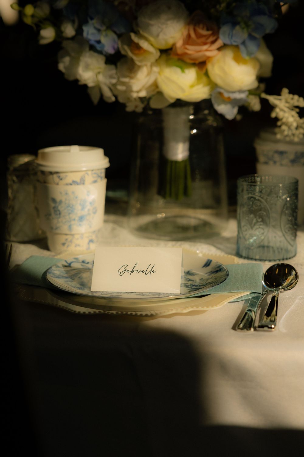 Elegant wedding place setting with blue floral china, name card reading Gabrielle, silver cutlery, and fresh flower bouquet centerpiece.