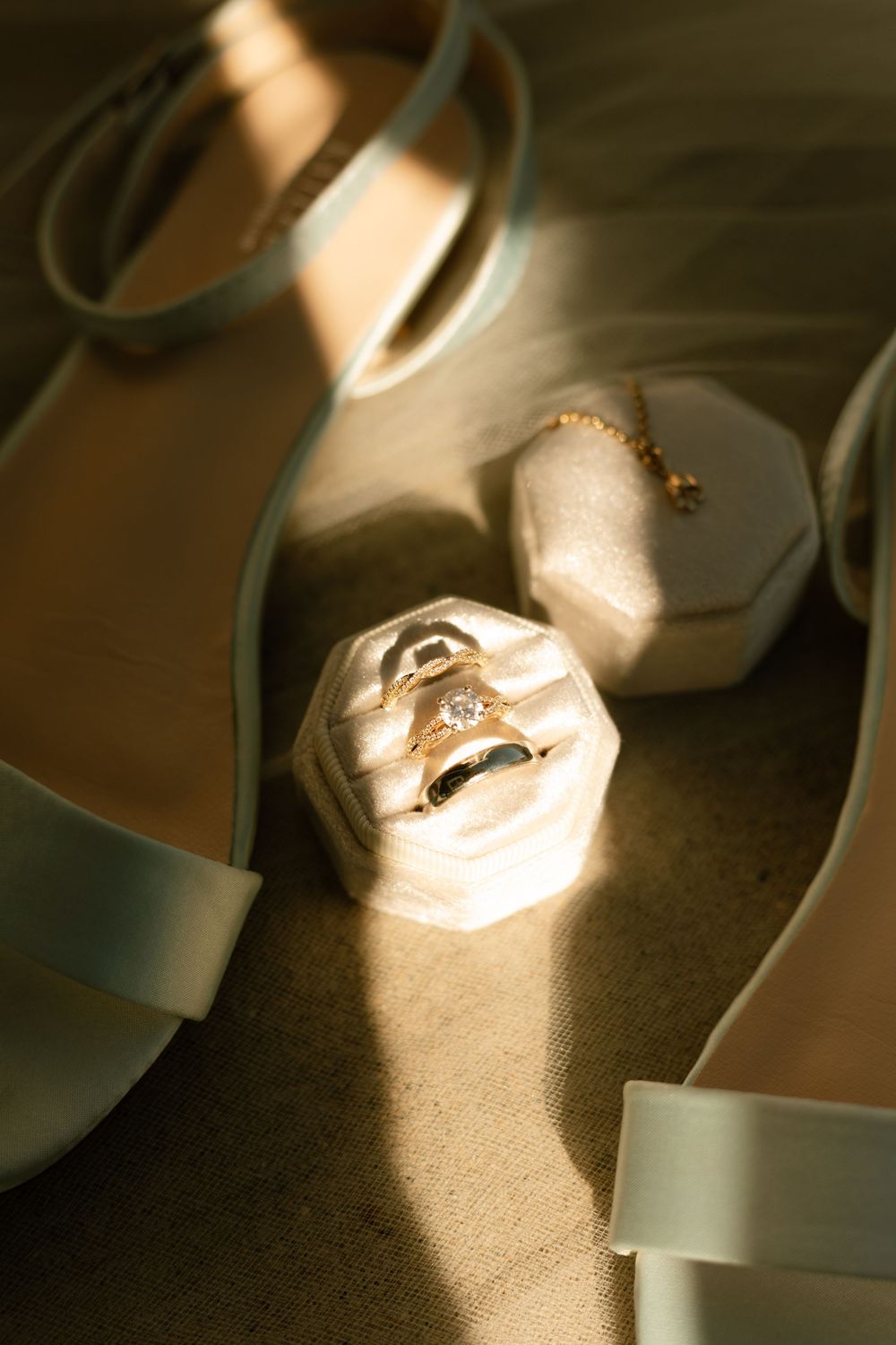 Wedding rings in a white velvet box beside elegant sage green heels, bathed in warm golden sunlight.