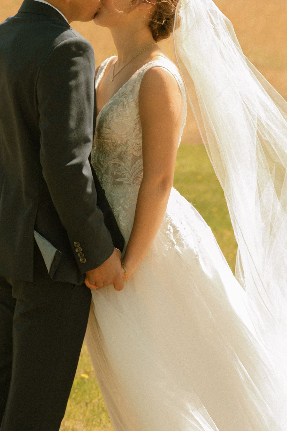 A bride and groom share a kiss while holding hands outdoors, the bride wearing a lace gown with flowing veil.