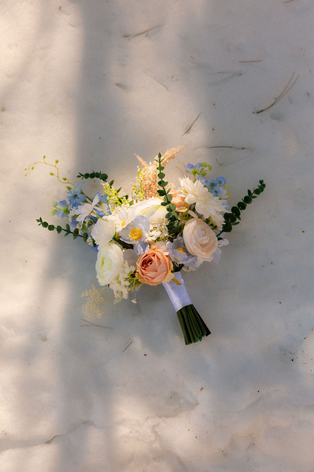 Elegant bridal bouquet with white, peach roses and greenery resting on sandy beach in natural sunlight.