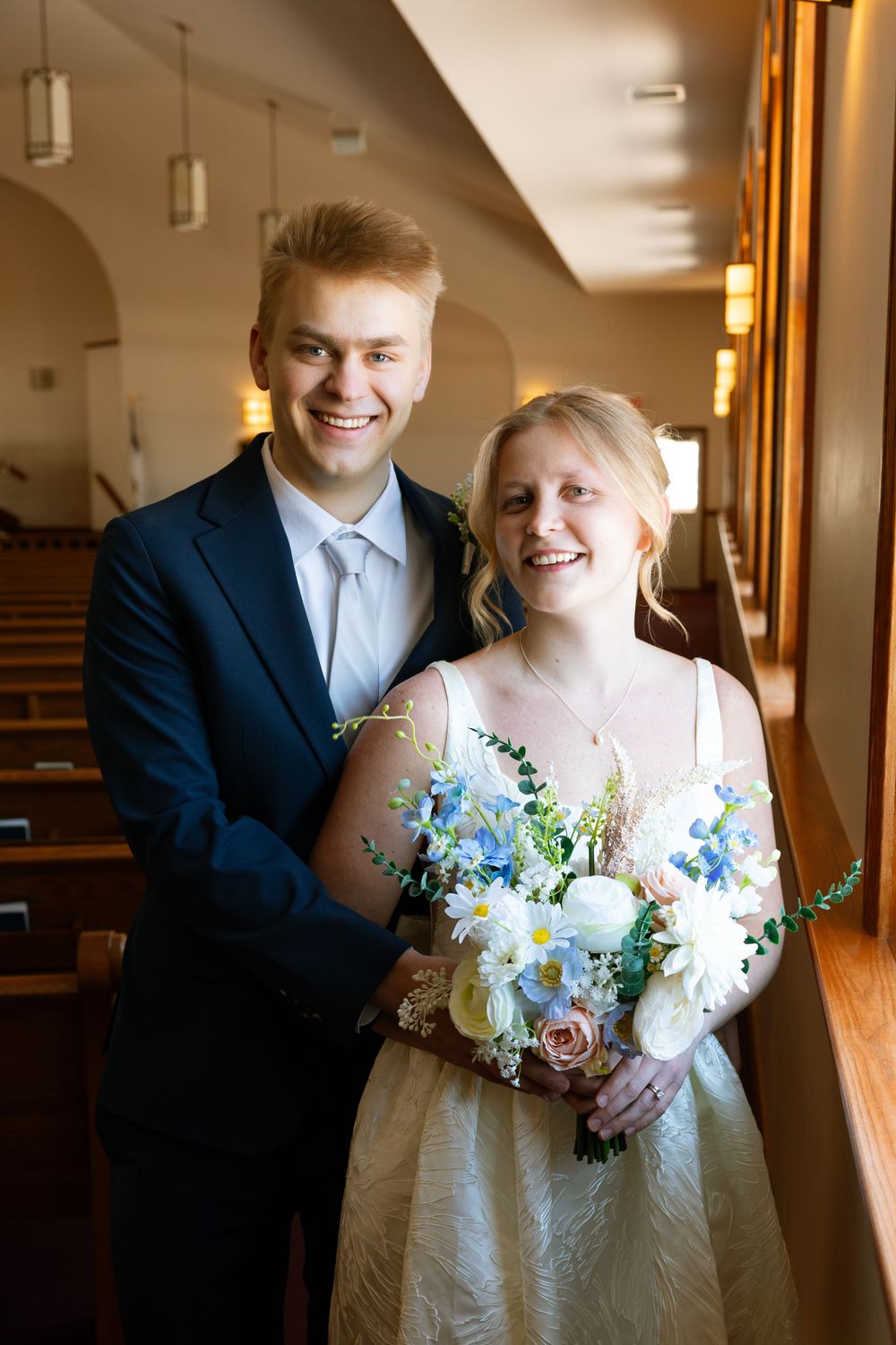 Happy newlyweds pose in a church aisle, bride holding blue and white bouquet, groom in navy suit smiling warmly.