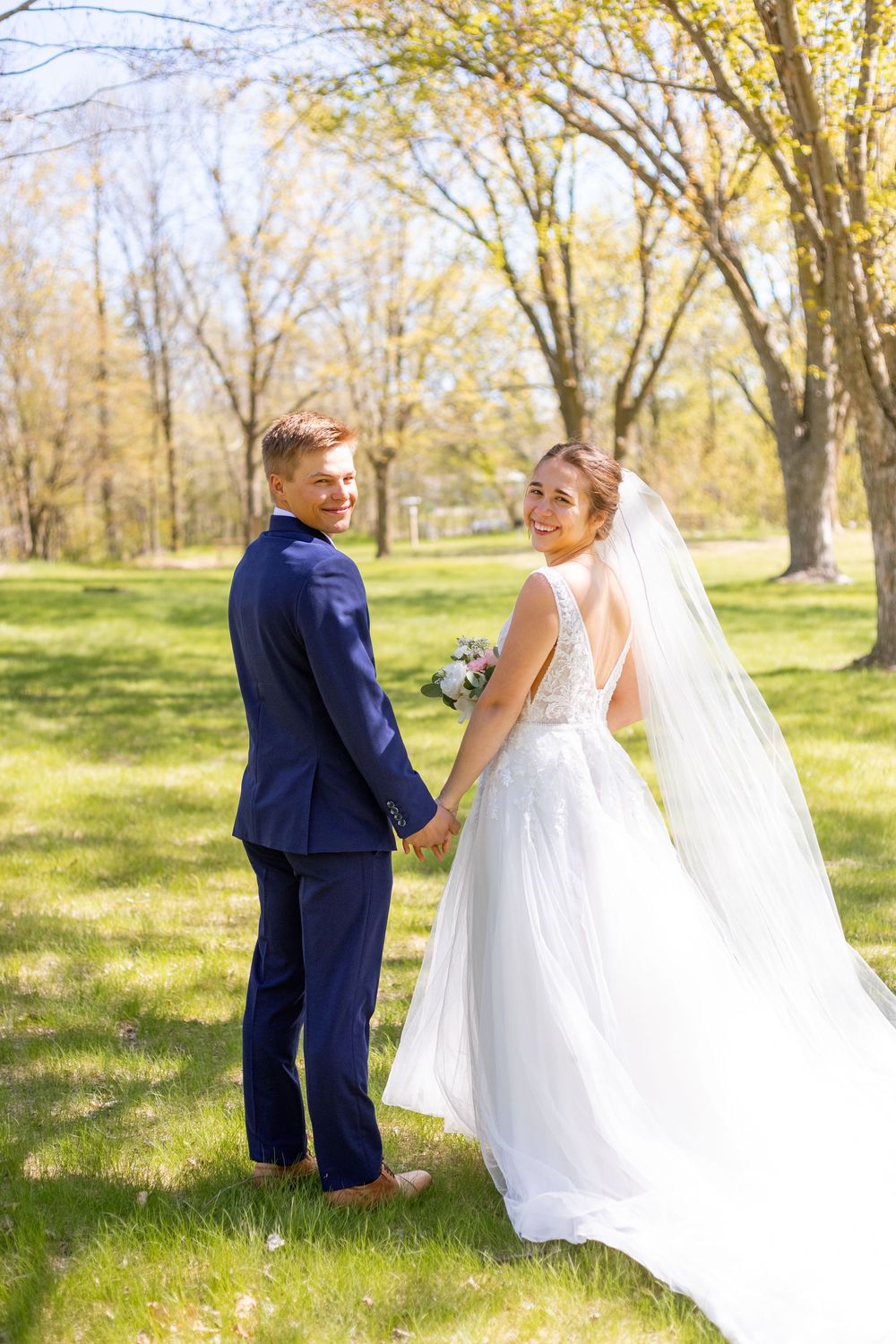 Smiling bride and groom holding hands in a sunny green park, bride in white gown with veil, groom in navy blue suit.