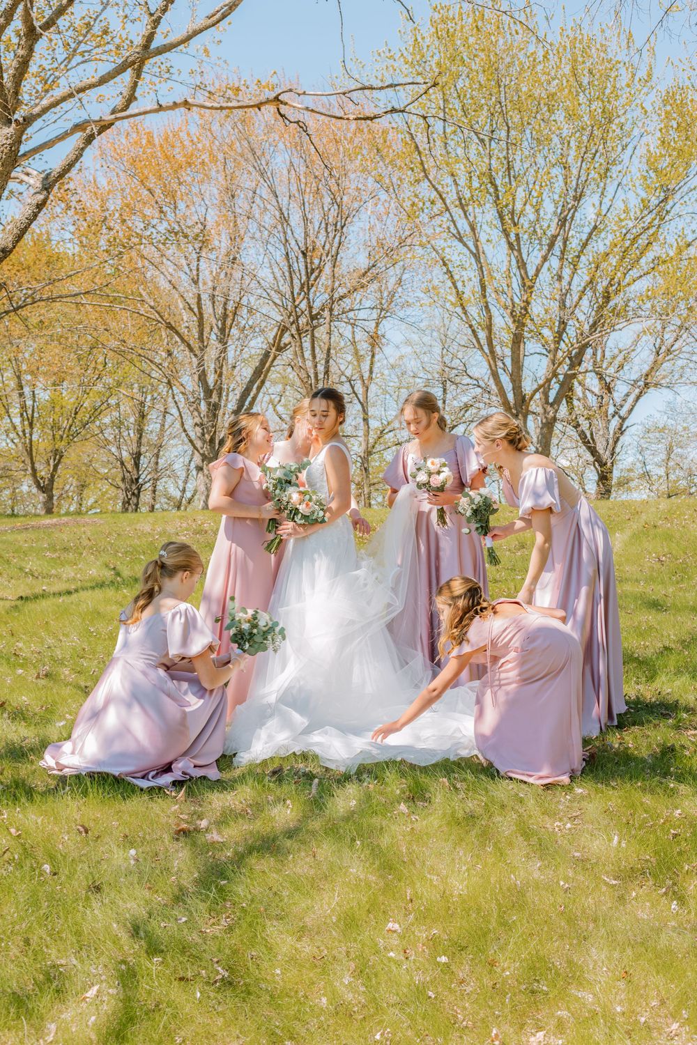 Bride in white gown with bridesmaids in pink dresses holding bouquets on a sunny green lawn.