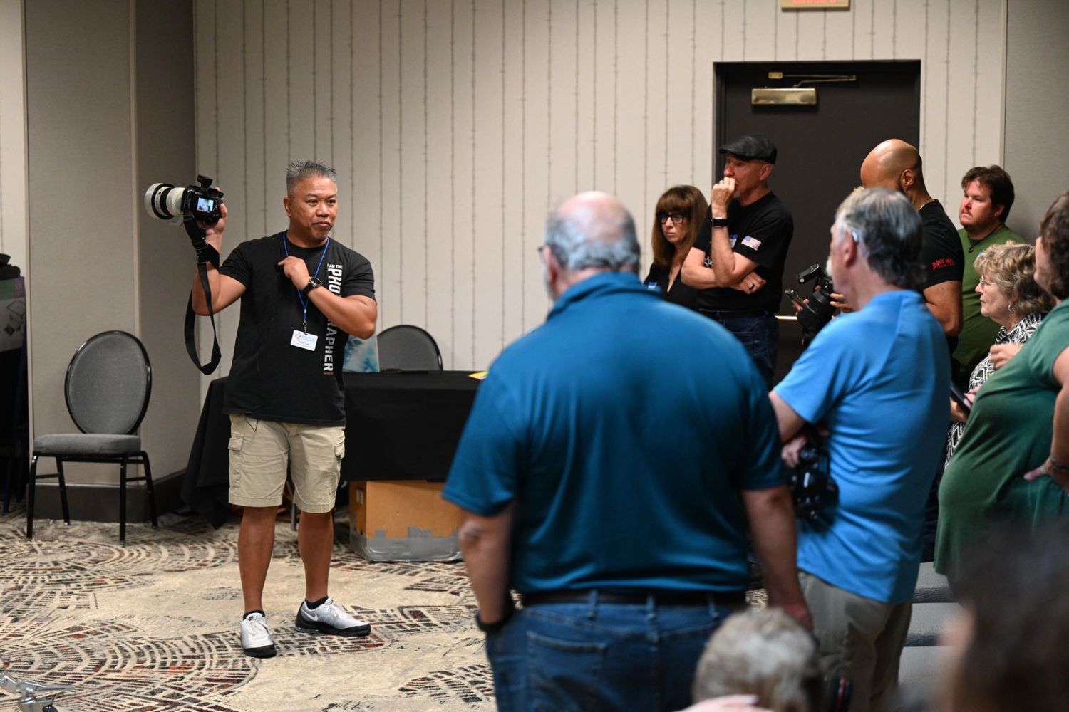 Photographer with camera addresses a crowd in a conference room during an indoor event or workshop.
